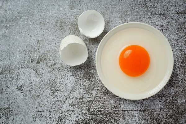 Quả trứng gà vỡ với lòng đỏ trong chén và hai nửa vỏ trứng bên cạnh trên nền màu xám.
